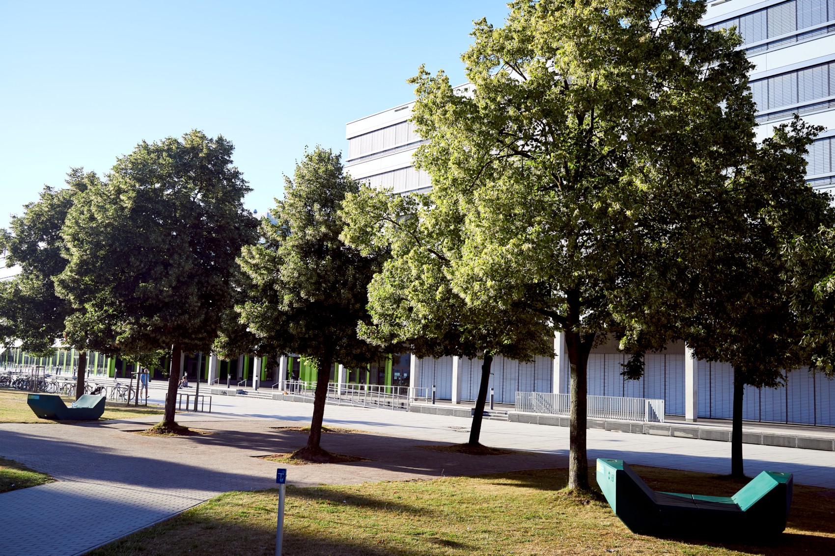 Das X-Gebäude der Universität Bielefeld wird teilweise von Bäumen verdeckt. Davor liegt eine Rasenfläche, mit einem Weg der zum X-Gebäude führt.